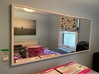 Full view of the large rectangular wall mirror hanging on a wall, reflecting a bedroom with a bed, window with Roman shade, and wall art.