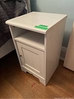 Front side view of one white veneer night stand showing cabinet door and open shelf, with some finish wear visible on edges and surface.
