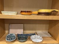 Two shelves showing variety of pottery pieces including small bowls, handled pans, a flat tray, and a picture frame wrapped in paper.