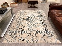 View of distressed style rug with beige and gray intricate motifs on floor between gray TV stand and brown leather couch.