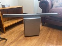 Soundbar placed on top of subwoofer near a chair and wooden shelf, showing shape and finish of items.