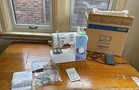 Brother sewing machine on wooden table near window with original box, foot pedal, instruction manuals, and small plastic accessory bags.