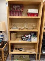 Full front view of wood veneer shelving unit containing various items such as glassware, dominoes, packaged goods, tools, and a lamp on adjacent wicker table.