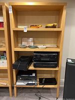 Front view of wooden veneer shelving unit showing four shelves holding electronic equipment and decorative ceramic dishes.