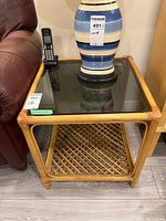 Square bamboo framed side table with dark glass top and lower lattice bamboo shelf beside leather chair and wooden cabinet. A telephone and ceramic lamp are visible on top.