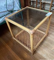 View of the entire side table showing smoked glass top, rattan cane frame, and lower woven cane tier on wooden floor.