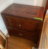 Wood side table showing geometric inlay pattern on the top surface, two drawers with distinct metal handles, and rich brown wood finish. Minor finish flaws visible.