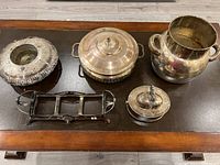 Five tarnished silver plate serving pieces displayed on a wooden table: a bowl, two lidded dishes, a large vessel, and a holder tray.