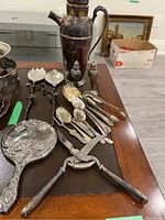 Silver plated seltzer jug, cutlery including forks, spoons, salad spoons, meat scissors, ladle, and assorted smaller pieces all tarnished, laid out on wood surface.