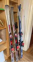 Photo showing two pairs of skis standing upright, Fisher skis in blue and white, Atomic skis in silver with red bindings, and a pair of black ski poles behind skis.