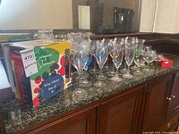 Wide shot of all stemware lined up on a wooden cabinet including boxed set of wine glasses, multiple champagne flutes, and a glass pitcher.