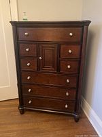 Front view of a dark wooden tallboy dresser with seven drawers and a small center cabinet door with silver round knobs.