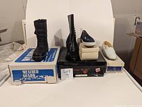 Four pairs of vintage shoes and boots displayed on their boxes including a pair of black weather guard winter boots, black leather lace-up boots, blue flats, and white loafers.