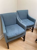 Photo showing two blue upholstered armchairs with wooden legs placed on a wood floor, corner of room visible.