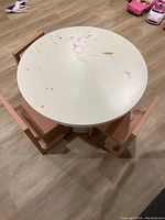 Top view of the round white kids table with clear visible paint splatters and marks on surface, surrounded by three pink wooden chairs on a laminate floor.