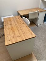 Two IKEA desks with natural wood tops and white drawer bases, one with five drawers, the other with a cabinet, placed next to each other with two white leather chairs in front.
