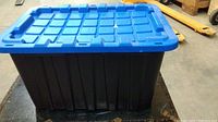 Front view of a black rectangular plastic storage bin with a blue lid that has a grid pattern and latch points.