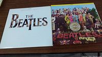 Photo of two Beatles vinyl albums placed side by side on a wooden surface showing cover art for '20 Greatest Hits' and 'Sgt. Pepper's Lonely Hearts Club Band'.