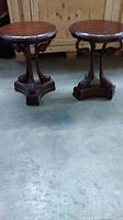 Pair of medium brown round wood end tables with ornate carved bases shown side by side on concrete floor against wood backdrop.