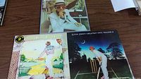 Three Elton John albums laid out on table: Yellow Brick Road with cover torn on bottom, Elton John self-titled album, and Greatest Hits Volume II.