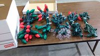 Five strands of large bulb Christmas lights laid out on a table beside an empty cardboard box showing three strands with bulbs and two without or partial bulbs.