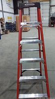 Full view of Werner 6-foot metal step ladder shows orange side rails, silver steps, black plastic top with 'Werner' logo in warehouse setting.