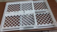 Full view of white plastic adjustable baby gate laid out flat on wood surface showing diamond-shaped perforations and sliding adjustment mechanism.