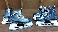 Two pairs of Bauer men's ice skates shown side by side on a wooden floor, grey with black accents, visible wear and marks on boots and blades.