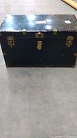 Front and top view of black vintage Monarch trunk showing metal latches and central lock plate, surface wear, missing key.