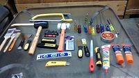 Full spread of hand tools laid out on a glass surface including hammer, paintbrushes, screwdrivers, utility knife, ear plugs, adhesive tubes and saw.