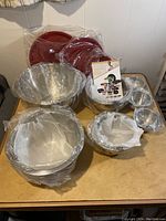 Overview of 14-piece Wolfgang Puck stainless steel bowl set showing multiple bowls and red lids wrapped in protective packaging on wooden table.