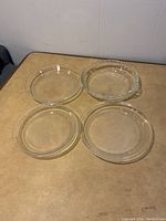 Four clear glass pie plates displayed on a table, three with plain edges and one with decorative edge and handles.