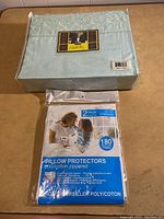 New packaged double sheet set in light blue with embroidered floral pattern on packaging label and a new sealed two-pack of white poly/cotton zippered pillow protectors with 180 thread count percale stated on package.