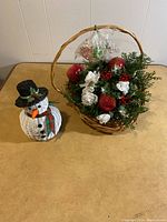 Full view of the Christmas basket with artificial greenery and decorations next to the white wicker snowman figurine.