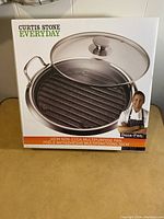 Front view of the Curtis Stone Everyday 30 cm non-stick multipurpose pan box showing pan with grill ridges and glass lid.