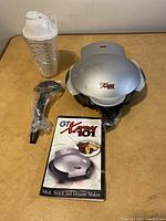 Closed GT Xpress 101 cooker with cookbook, measuring cup, and power cord on table.