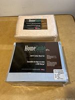 Two sealed HomeSuite Collection bedding sets on a surface, one in pale peach with leaf designs and one in solid gray, both labeled 500TC cotton sheets for double beds.