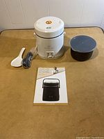Complete lot including Wolfgang Puck portable rice cooker, extra pot and lid, rice paddle, power cord, and user manual on beige table