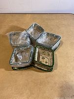 Six square small dishes stacked in two groups, showing the green and white floral leaf patterned rims and interiors with some dishes wrapped in plastic.