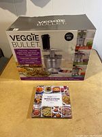 Box of Veggie Bullet food processor set and Whole Food Nutrition recipe book on a table.