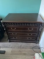 Front view of the file cabinet showing rich brown finish and ornate carving along the top edge with three drawers each with metal handles.