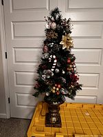 Mini artificial Christmas tree decorated with ornaments including pine cones, candy cane, gold poinsettia, and baubles, standing on ornate base.