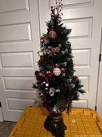 Mini artificial Christmas tree decorated with pine cones, baubles, berries, and other ornaments, standing in a gold pot base against a white door background.