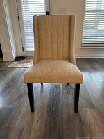 Front view of the beige fabric upholstered Parsons style dining chair with vertical stitched channels and black legs on wood floor.