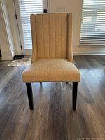 Front view of beige fabric upholstered Parsons dining chair with vertical channel tufted backrest, padded seat, and black legs on wood floor.