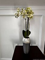 Tall hexagonal ceramic vase holding a plastic orchid plant with white, pale yellow flowers and green leaves.