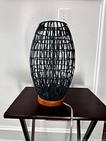Side view of the black woven oval-shaped shade lamp on a wooden base with power cord visible on a table.