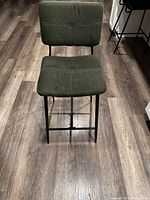 Front view of upholstered army green counter stool showing cushioned seat and backrest with stitched square pattern and black metal frame with footrest