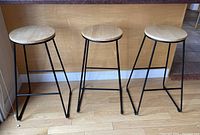 Three bar stools with round wooden seats and black metal legs arranged side by side on a wood floor.
