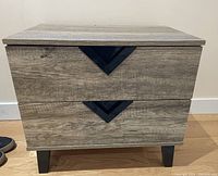 Front view of bedside table showing two drawers with black V-shaped handles and wood grain laminate finish.